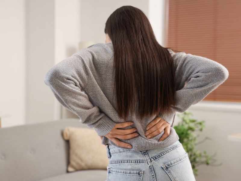 Mujer de espaldas sujetando su zona lumbar con ambas manos, expresando una molestia física común derivada de la postura al trabajar.