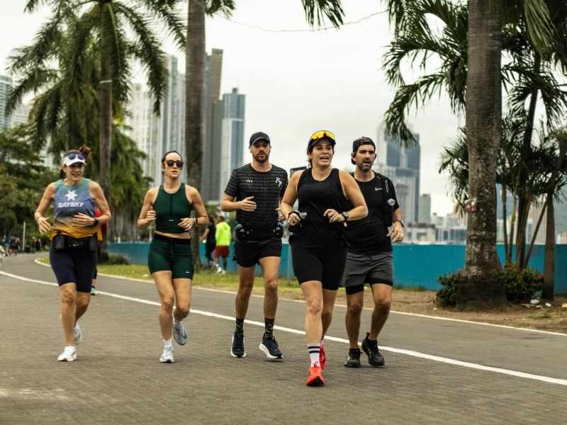Grupo diverso de personas corriendo juntas en una vía urbana cerca del mar, destacando la importancia de la actividad física compartida y la constancia en el deporte
